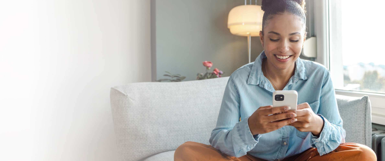 A Black woman in a blue jean button-up shirt is sitting cross-legged on the couch, smiling while using her smartphone.